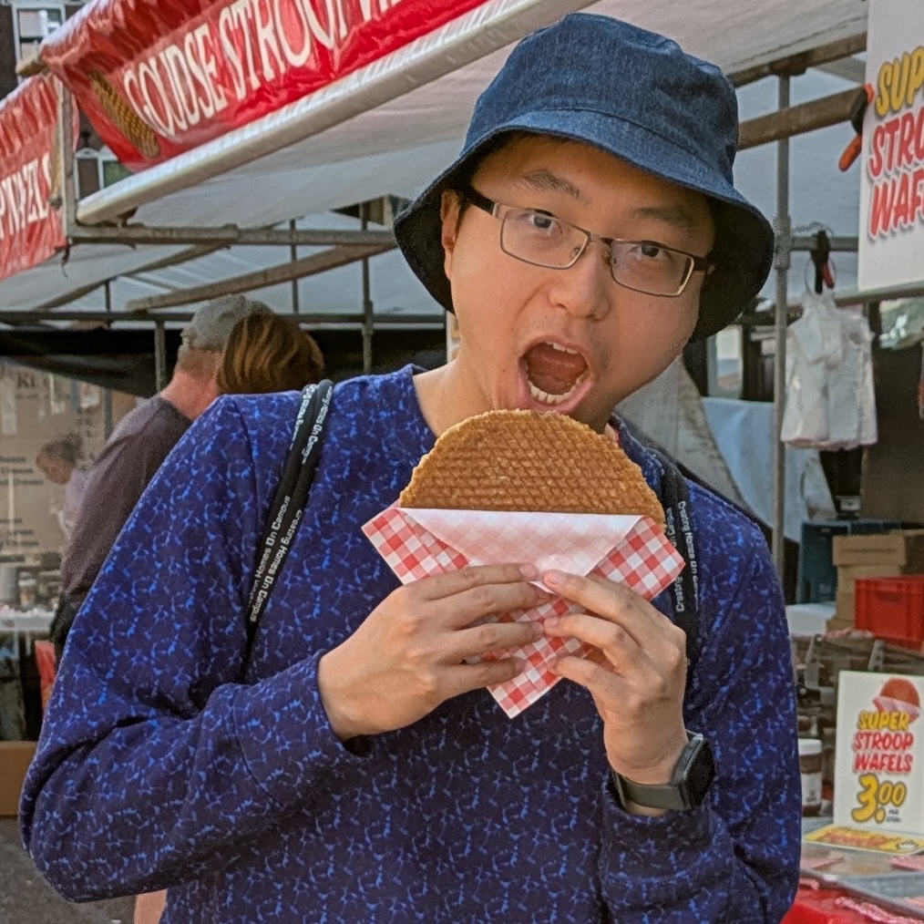 An Asian man in a bucket hat and glasses about to eat a large stroopwafel.